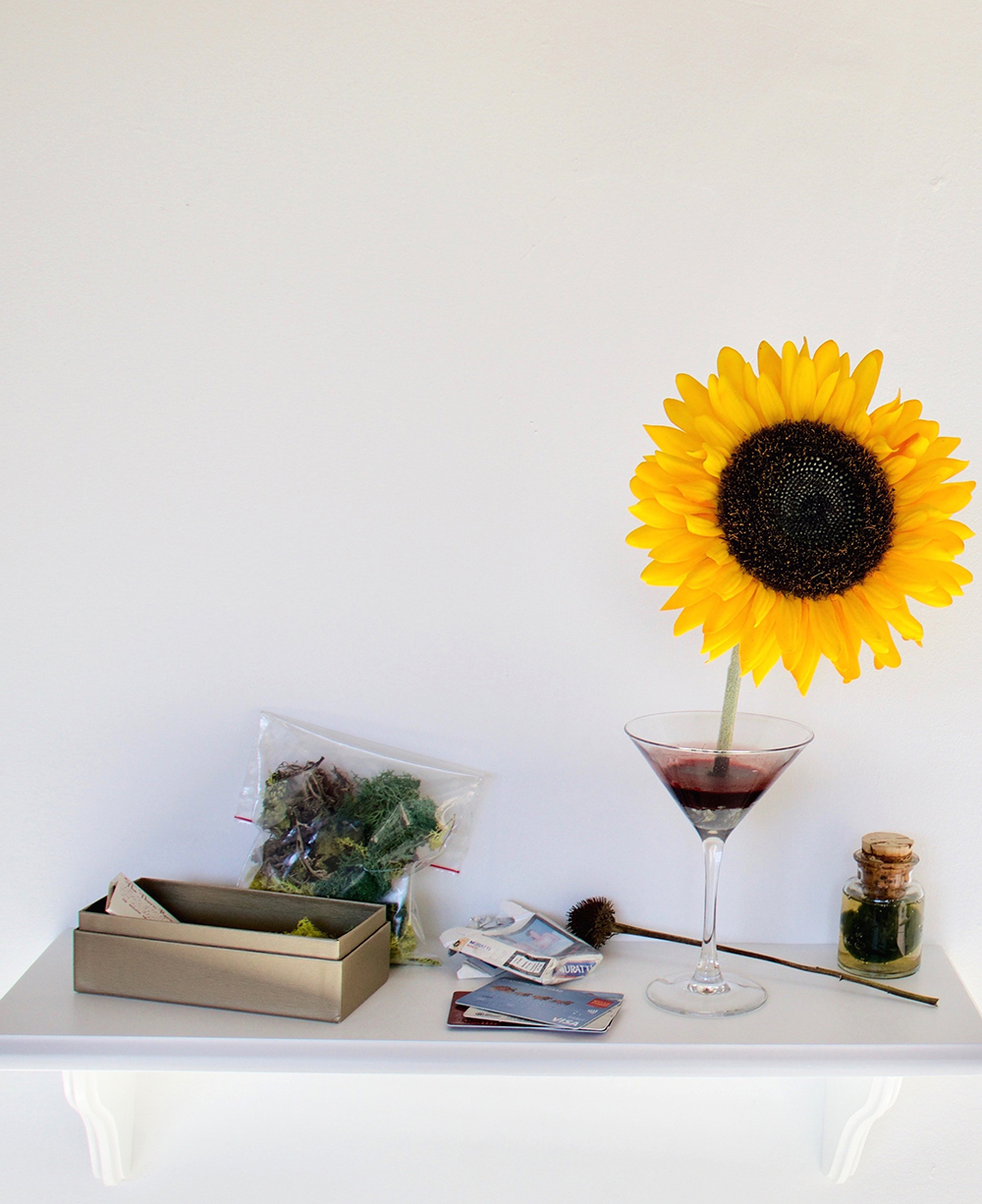 A shelf with a bloody martini glass that has a flower growing out of it along with other small ritual objects